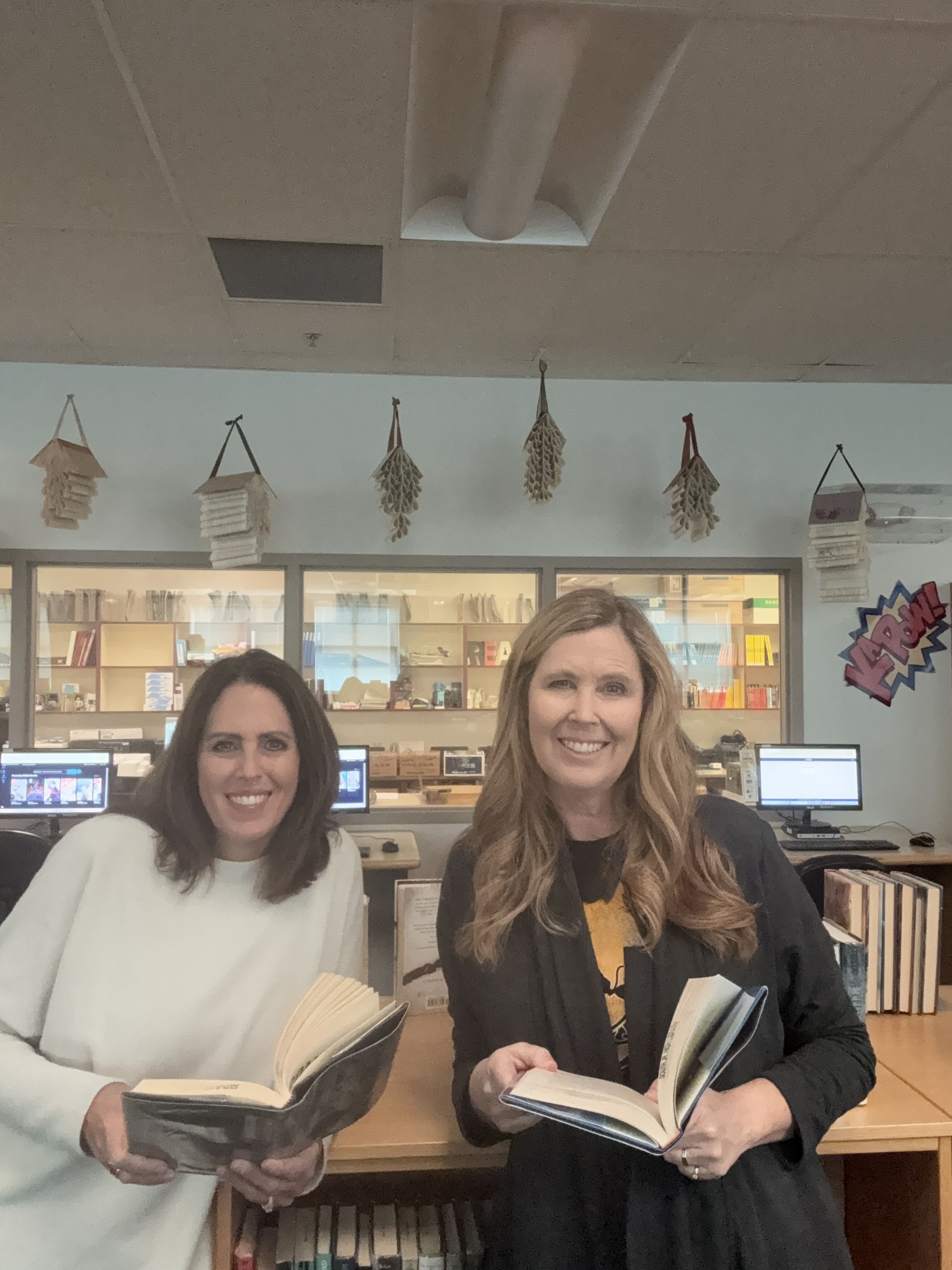 Two smiling women stand in a library, each holding open books to show the camera.